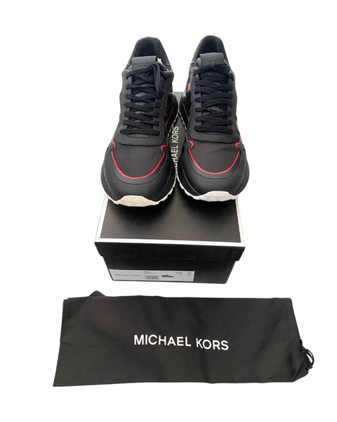 Black sneakers with red accents on a black box, accompanied by a Michael Kors pouch.