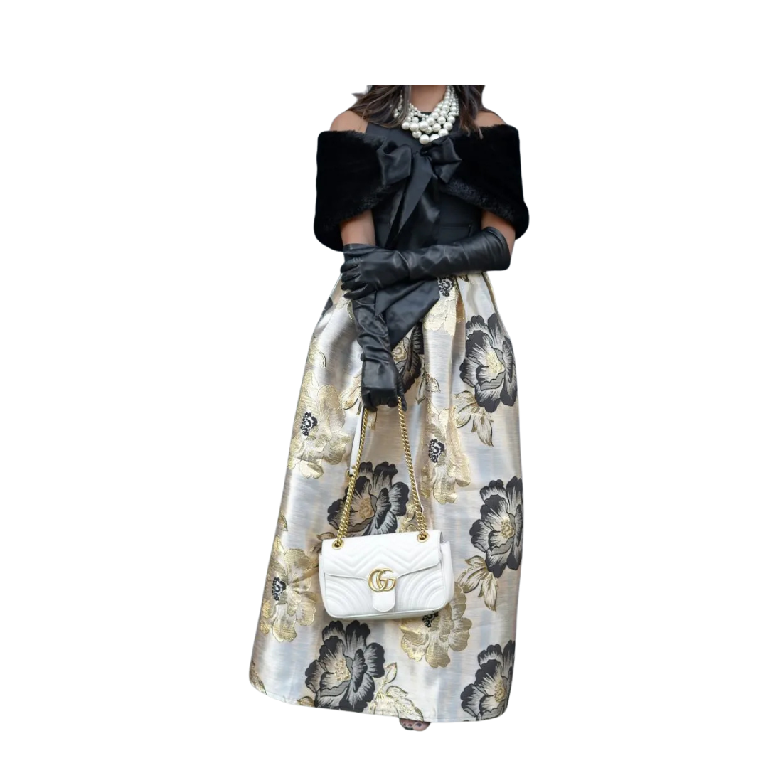 Woman wearing a floral skirt, black top, and white gloves with a white handbag on a white background